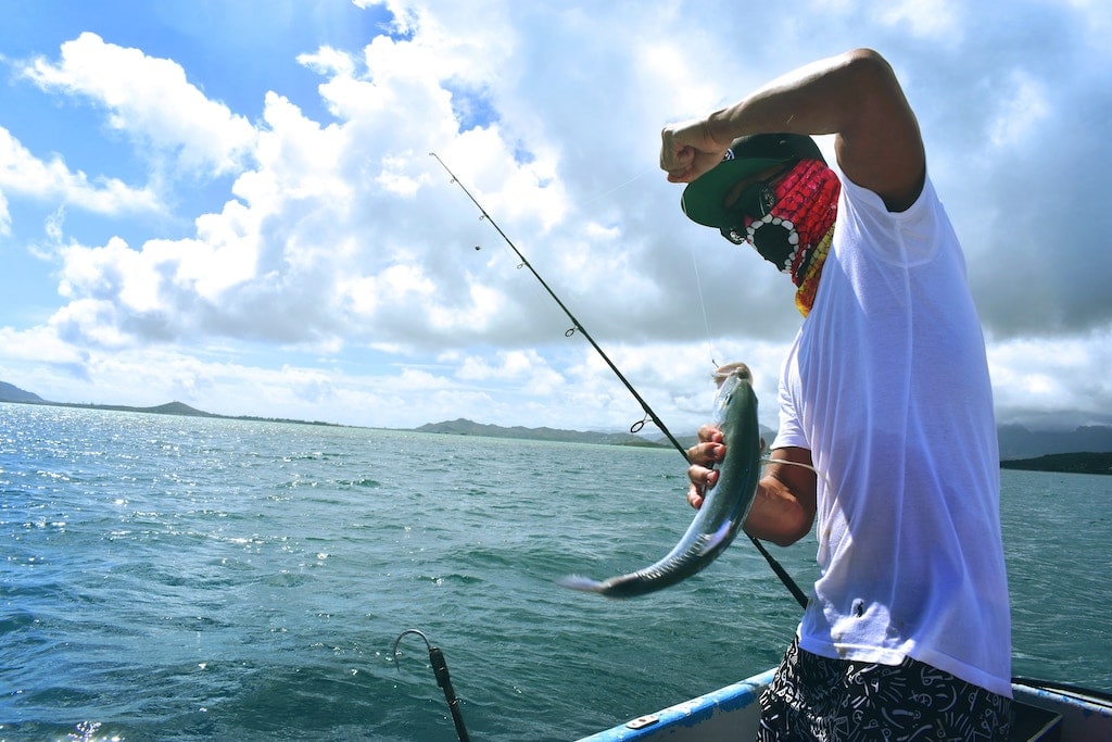 A man wearing a face mask reels in a fish while on a fishing boat.