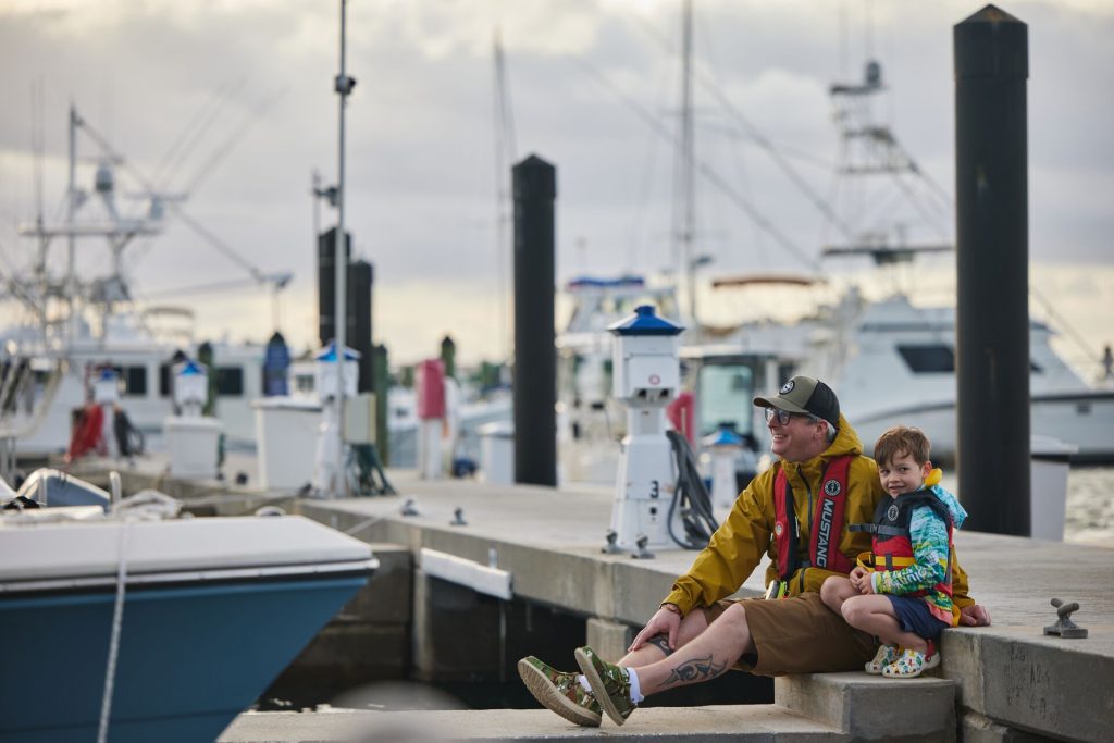 A man and boy sit on the dock at a marina.