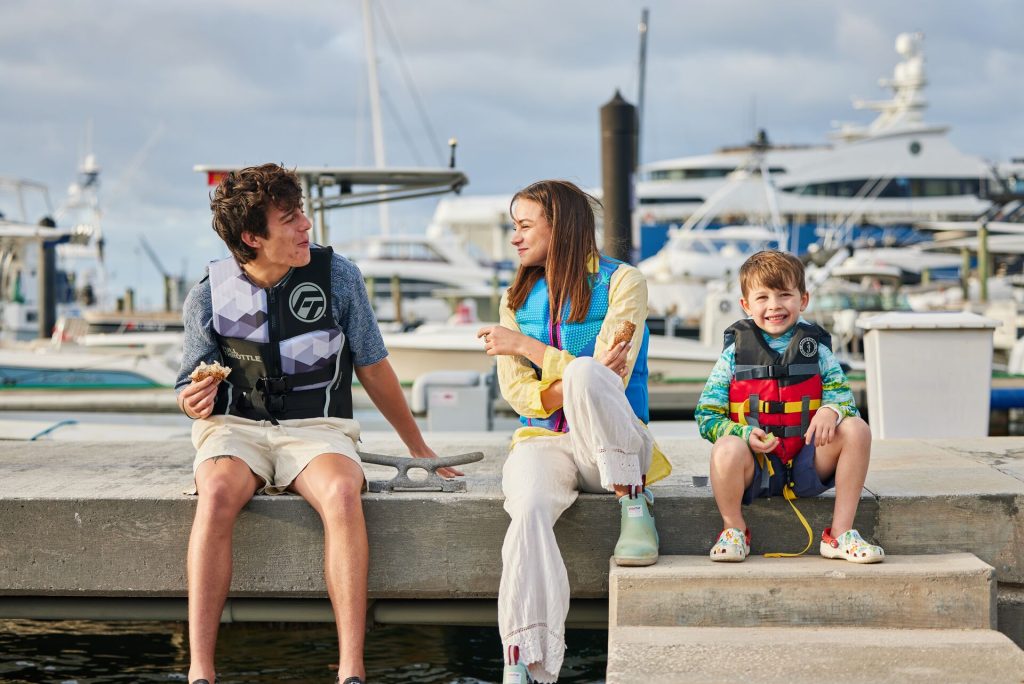 Three kids sit on a dock wearing life jackets, represent how to buy children's PFDs.