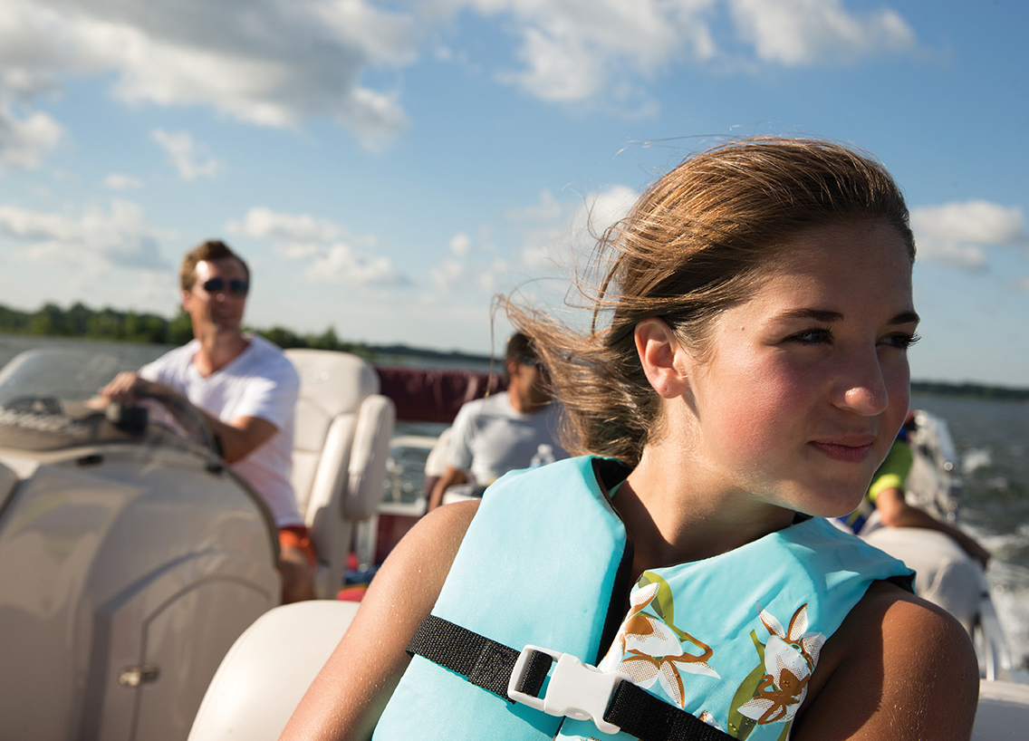 girl sitting in boat with lifejacket on and father in background