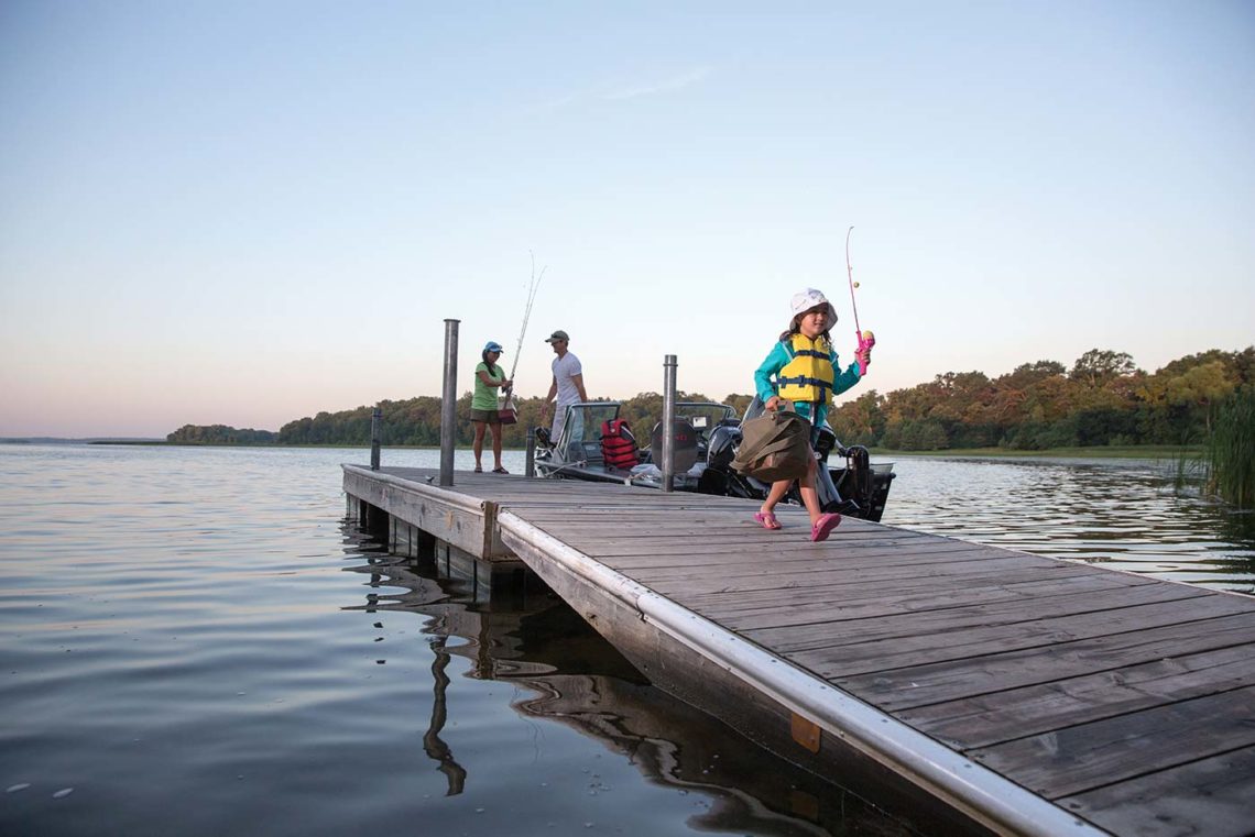 Girl carrying tackle box on dock with parents in background