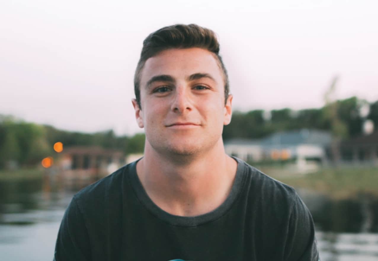 A young man stands smiling in front of a blurred background of a lake and boats